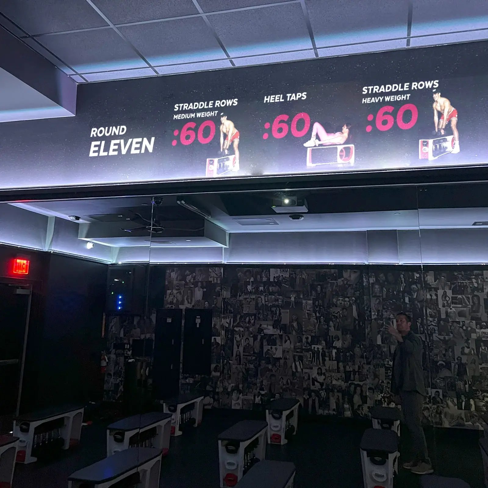 Wide studio display showing a workout round with straddle rows, heel taps, and interval timers above the training floor